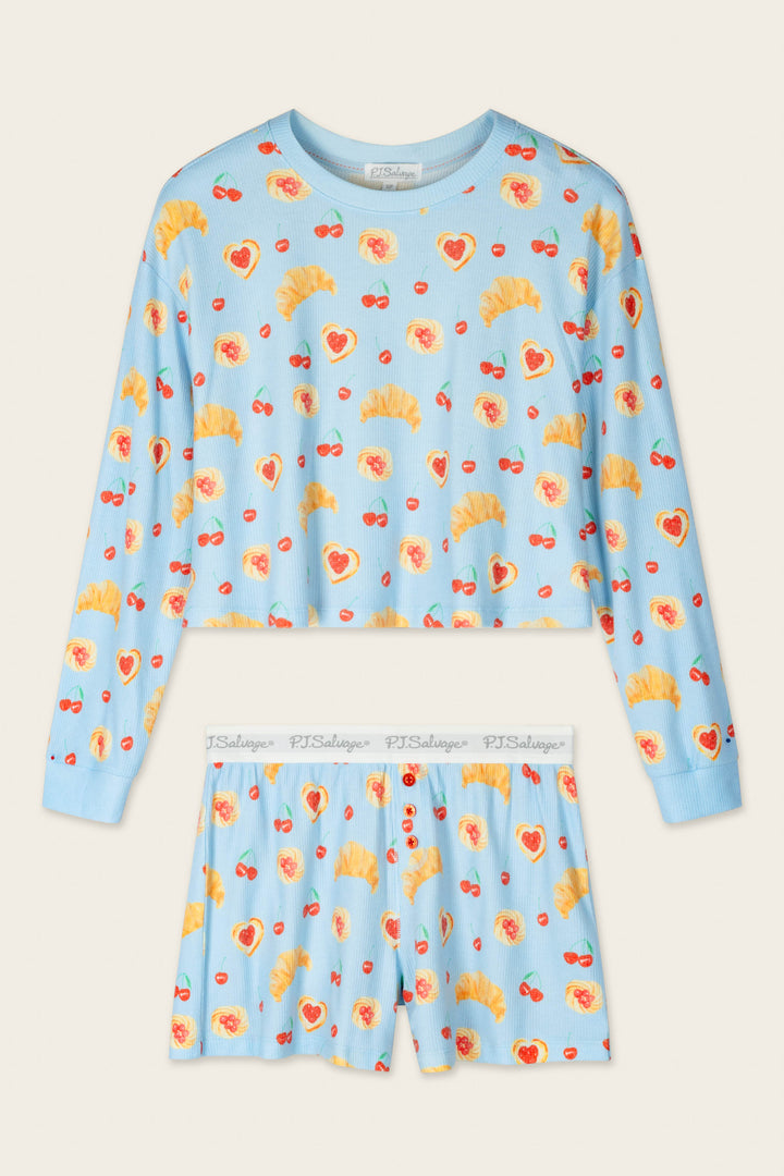 The P.J. Salvage Boyfriend Boxer Pastry Print Long Sleeve & Shorts PJ Set features a light blue long-sleeve top and shorts decorated with playful prints of croissants, hearts, and cherries. The shorts include a white elastic waistband printed with "PJ Salvage." The set is laid flat on a beige background.