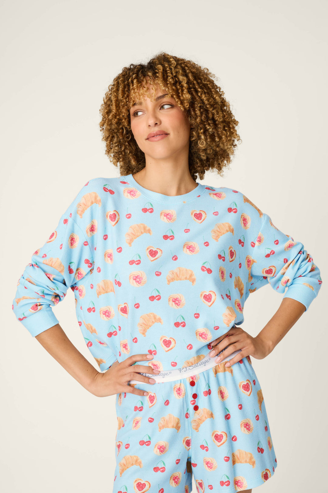 A person with curly hair wears the P.J. Salvage Boyfriend Boxer Pastry Print Long Sleeve & Shorts PJ Set, featuring a blue design decorated with croissants, hearts, and sunglasses. They stand with hands on hips, looking off to the side and smiling slightly.