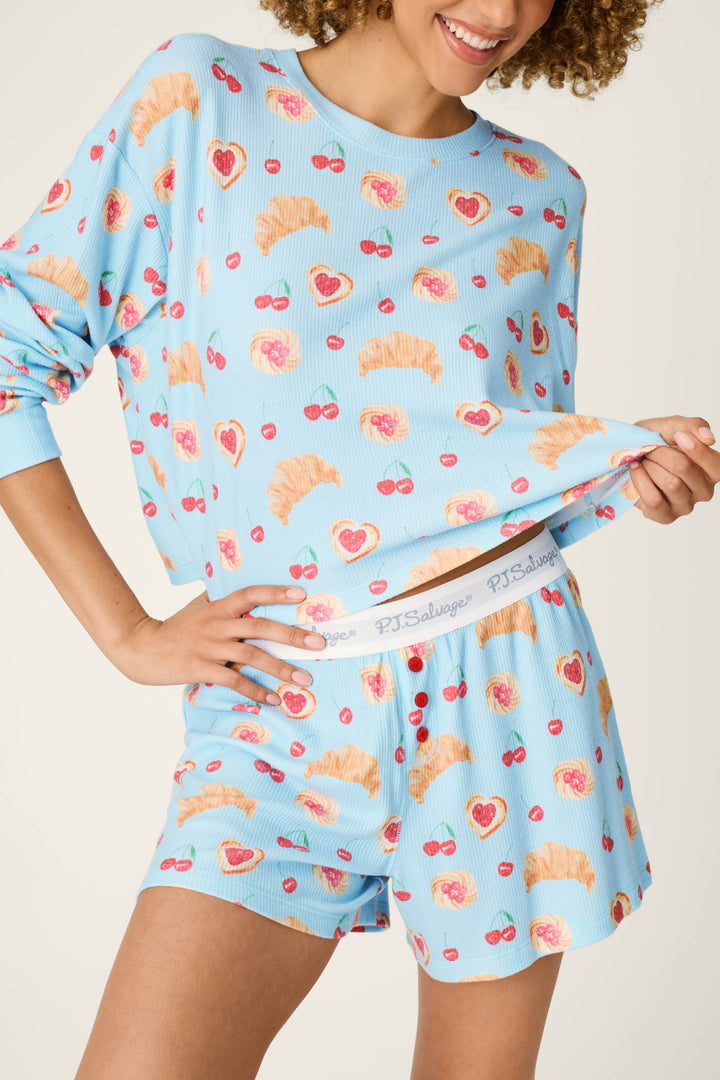 A person models the P.J. Salvage Boyfriend Boxer Pastry Print Long Sleeve & Shorts PJ Set, featuring a matching light blue design with croissant, cherry, and strawberry motifs. The individual lifts the shirt to reveal a waistband printed with the P.J. Salvage brand name; only their lower face and body are visible.