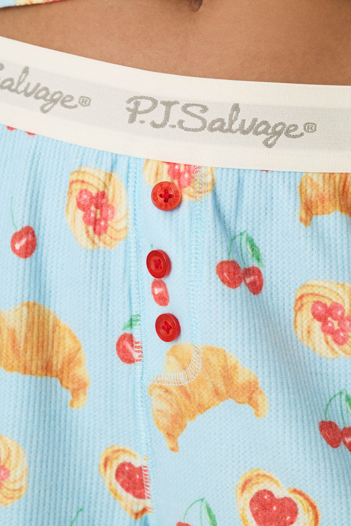 Close-up of blue pajama shorts from the P.J. Salvage Boyfriend Boxer Pastry Print Long Sleeve & Shorts PJ Set, featuring a croissant and cherry print, red buttons, and a white elastic waistband.