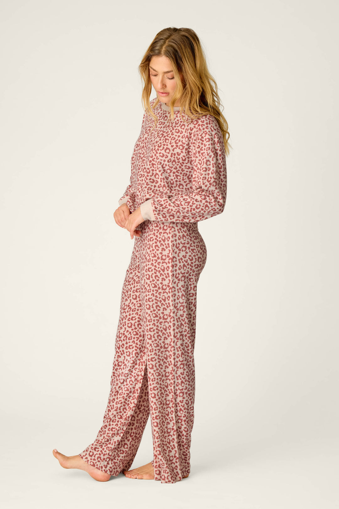 A woman with long, loose hair stands barefoot against a plain background, looking down as she adjusts her sleeve while wearing the P.J. Salvage Boyfriend Boxer Leopard Print Long Sleeve PJ Set.