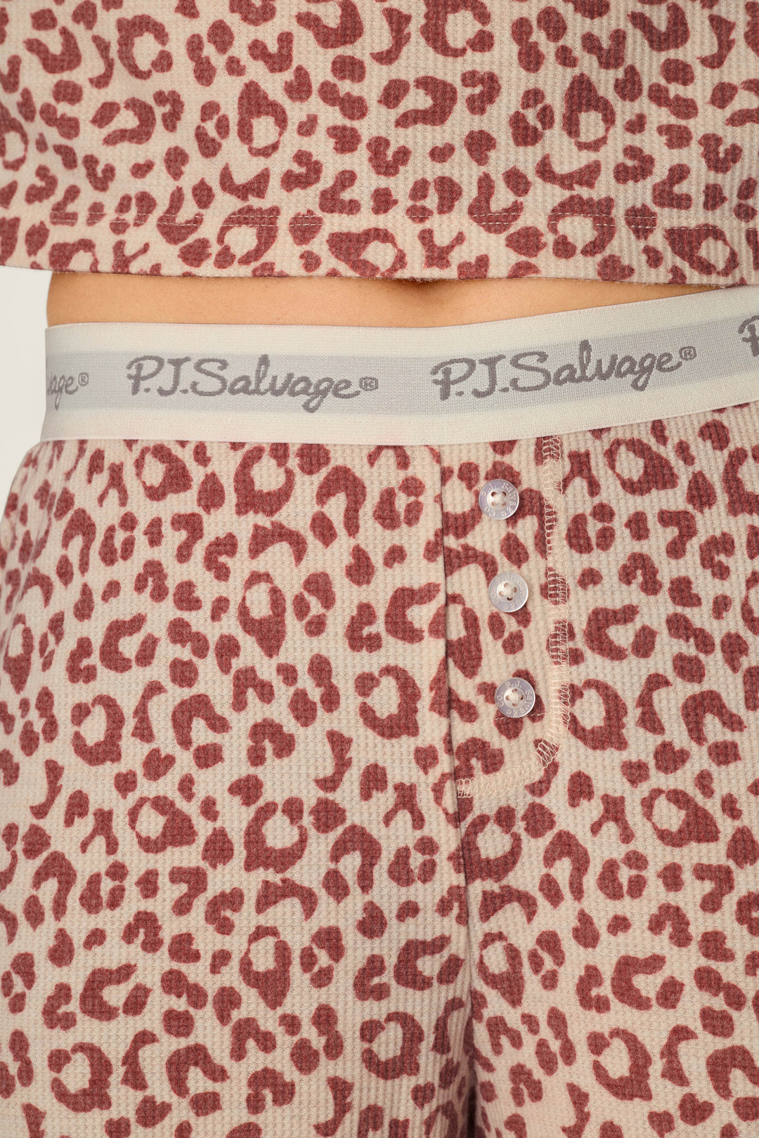 Here’s a rewritten version using your product data: Close-up of a person wearing the P.J. Salvage Boyfriend Boxer Leopard Print Long Sleeve PJ Set, featuring pink and red leopard print pajama pants with a ribbed texture, three buttons, and a gray waistband printed with the P.J. Salvage logo in cursive.