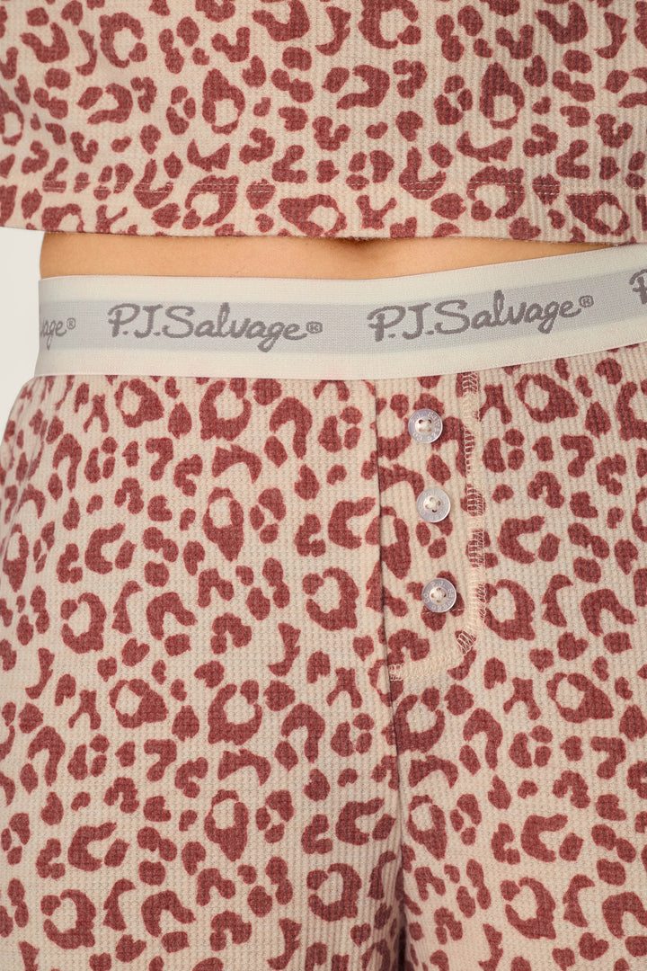 Here’s a rewritten version using your product data: Close-up of a person wearing the P.J. Salvage Boyfriend Boxer Leopard Print Long Sleeve PJ Set, featuring pink and red leopard print pajama pants with a ribbed texture, three buttons, and a gray waistband printed with the P.J. Salvage logo in cursive.