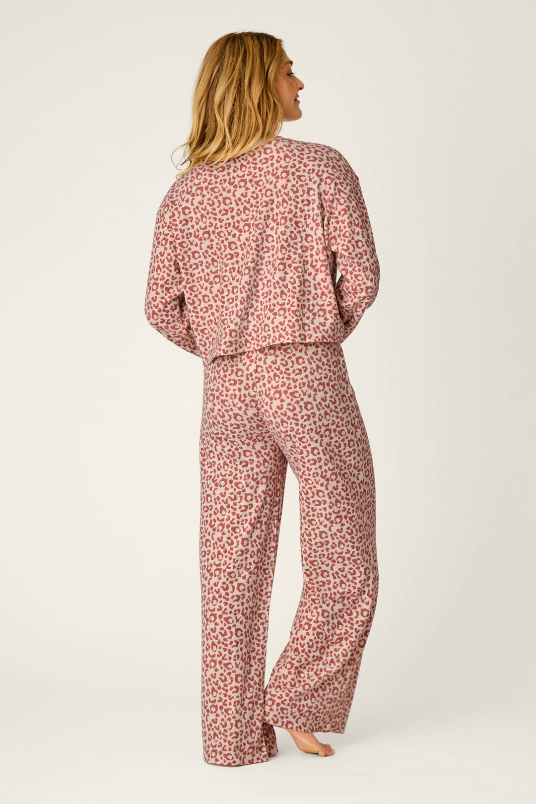 A woman stands barefoot facing away, wearing the Boyfriend Boxer Leopard Print Long Sleeve PJ Set by P.J. Salvage, featuring matching long-sleeve top and pants with a pink leopard print, set against a plain light background.