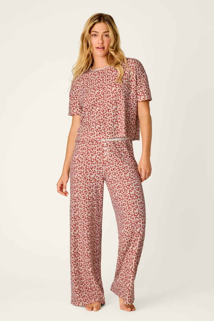 A woman stands barefoot against a plain light background wearing the P.J. Salvage Boyfriend Boxer Leopard Print Short Sleeve & Pants PJ Set, featuring a matching red leopard print with a short-sleeve top and wide-leg pants.