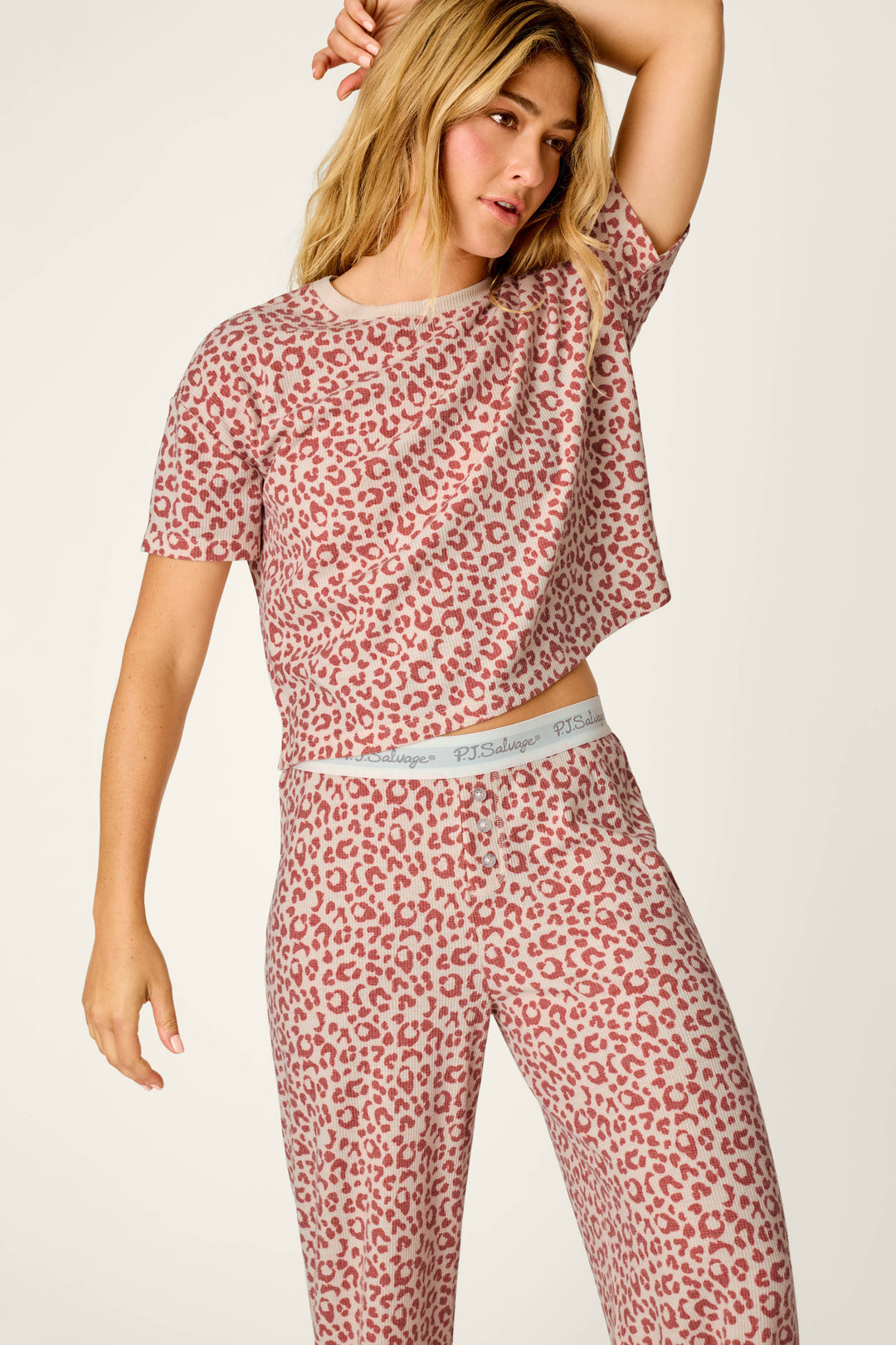 A person wearing the P.J. Salvage Boyfriend Boxer Leopard Print Short Sleeve & Pants PJ Set poses with one arm raised, looking to the side. The matching set features a pink and white leopard print, a short-sleeve top, and fitted pants with a branded waistband.