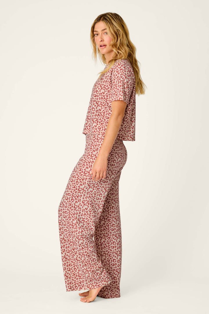 A woman stands barefoot in a side pose, wearing the P.J. Salvage Boyfriend Boxer Leopard Print Short Sleeve & Pants PJ Set, featuring a pink and red leopard print pattern, against a plain light background.