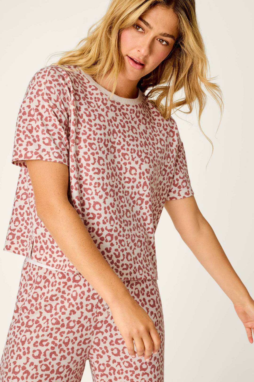 A woman with long blonde hair wears the P.J. Salvage Boyfriend Boxer Leopard Print Short Sleeve & Pants PJ Set, featuring a matching pink and white leopard print design, as she stands against a light background.