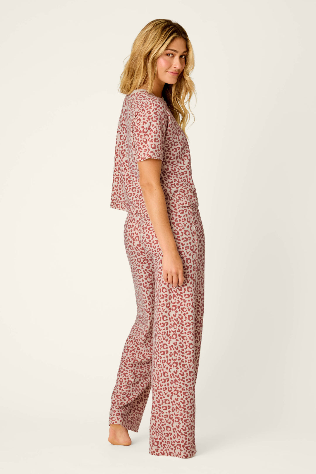 A woman stands in profile, barefoot against a plain light background, wearing the P.J. Salvage Boyfriend Boxer Leopard Print Short Sleeve & Pants PJ Set—a matching pink and gray leopard print pajama set featuring short sleeves and long pants. Her long, wavy blonde hair cascades down her back.