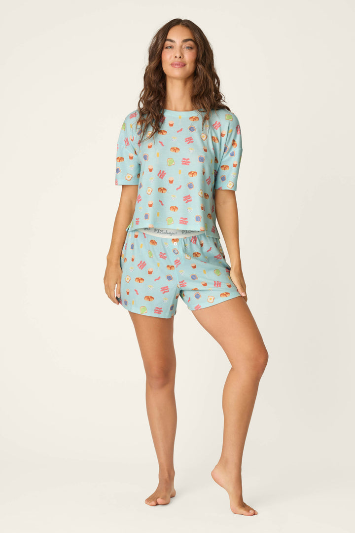 A woman with long, wavy brown hair stands barefoot, smiling softly in the P.J. Salvage Boyfriend Boxer Sunday Brunch Short PJ Set—a matching light blue pajama set with colorful food prints, featuring a short-sleeve top and shorts.