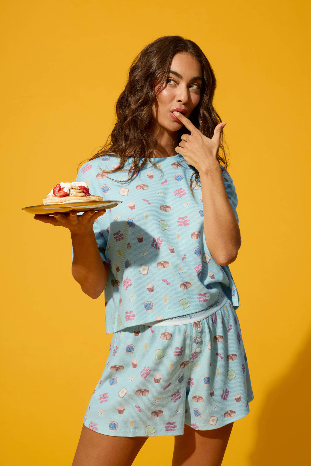 A woman in the P.J. Salvage Boyfriend Boxer Sunday Brunch Short PJ Set holds a plate of pancakes with strawberries and cream, playfully touching her finger to her lips against a bright yellow background.