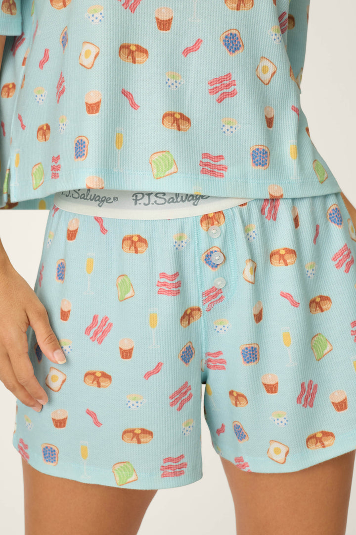 A person wears P.J. Salvage's Boyfriend Boxer Sunday Brunch Short PJ Set, featuring light blue pajamas with a breakfast food print. Only the lower torso and one hand are visible; "P.J. Salvage" is on the waistband.