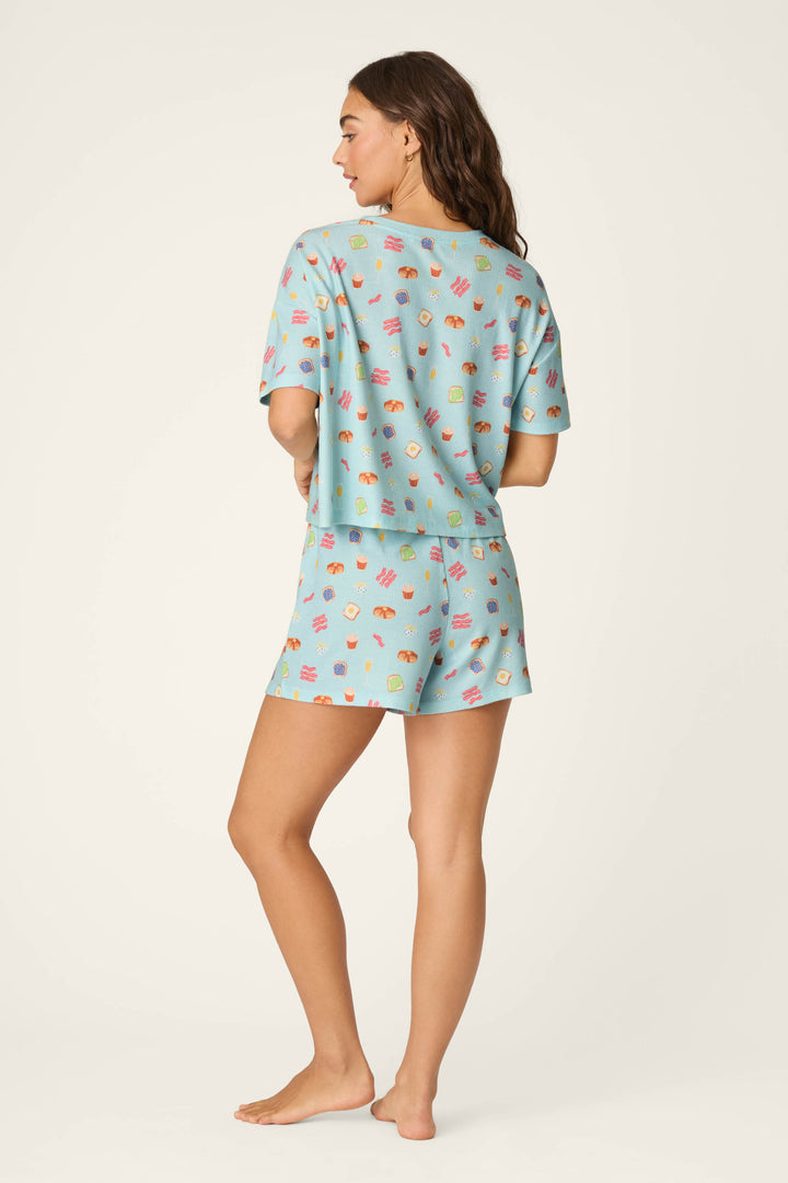 A woman stands barefoot facing away, wearing the P.J. Salvage Boyfriend Boxer Sunday Brunch Short PJ Set in light blue with colorful food patterns, against a plain light background.