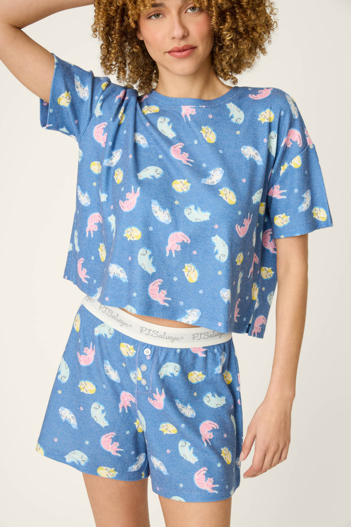 A person wears the P.J. Salvage Boyfriend Boxer Cat Nap Short PJ Set—blue with playful pink, yellow, and white fish prints. The set features a short-sleeved top and matching shorts with a white "P.J. Salvage" waistband.