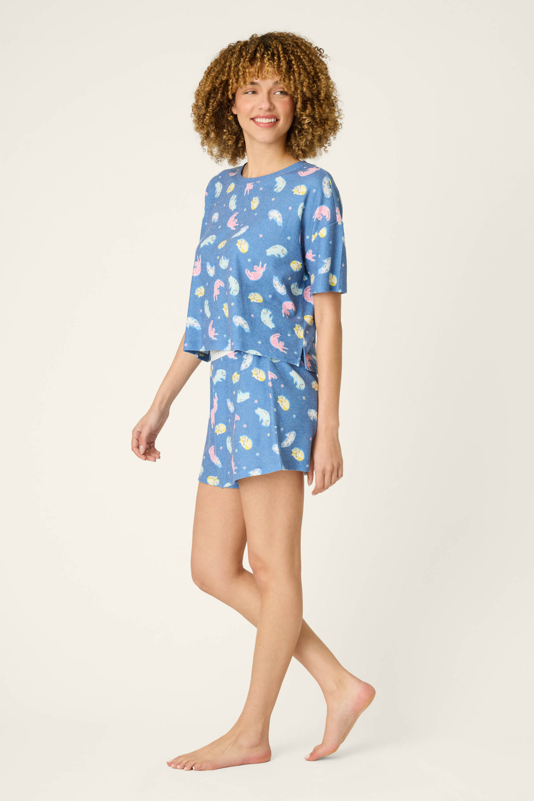 A woman with curly hair smiles barefoot in a P.J. Salvage Boyfriend Boxer Cat Nap Short PJ Set, featuring matching blue pajamas with playful prints, against a plain light background.