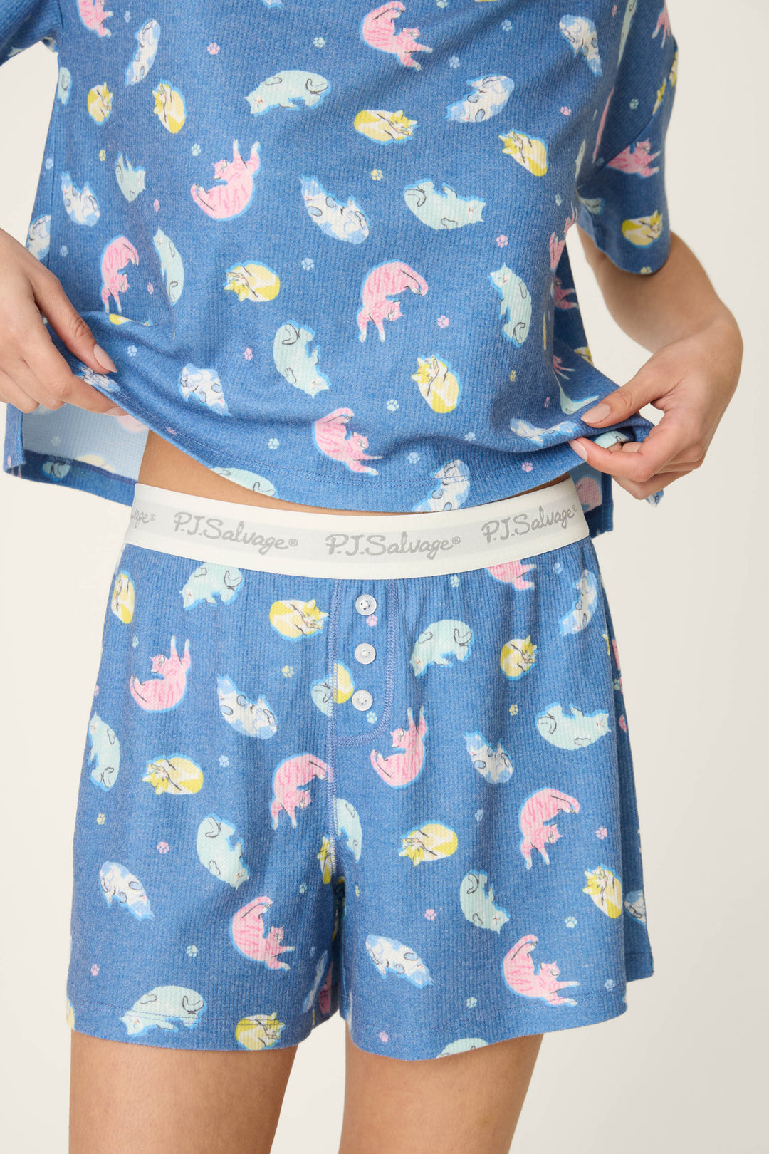 Someone wears the Boyfriend Boxer Cat Nap Short PJ Set by P.J. Salvage, featuring a blue background with colorful cat print and a slightly lifted top showing the P.J. Salvage-branded waistband of the shorts.