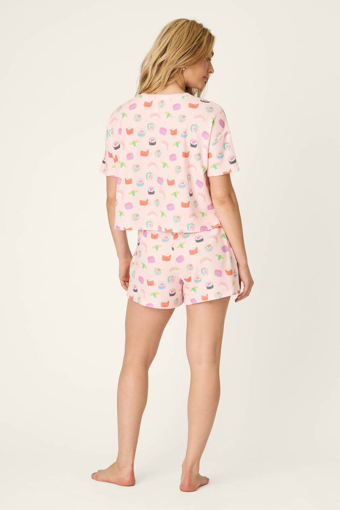 A woman with blonde hair stands barefoot, facing away, wearing the P.J. Salvage Boyfriend Boxer Sushi Bites Short PJ Set—a matching short-sleeve top and shorts with playful sushi print.