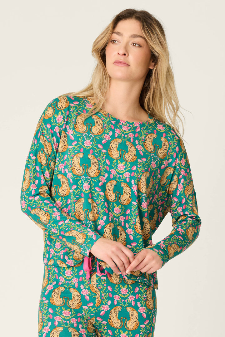 A woman wears the P.J. Salvage Cheetah Bonita Dreams Cheetah Print Long Sleeve Top, featuring a green base with an allover cheetah and pink floral pattern, standing with one hand at her waist and gazing slightly to the side against a plain light background.