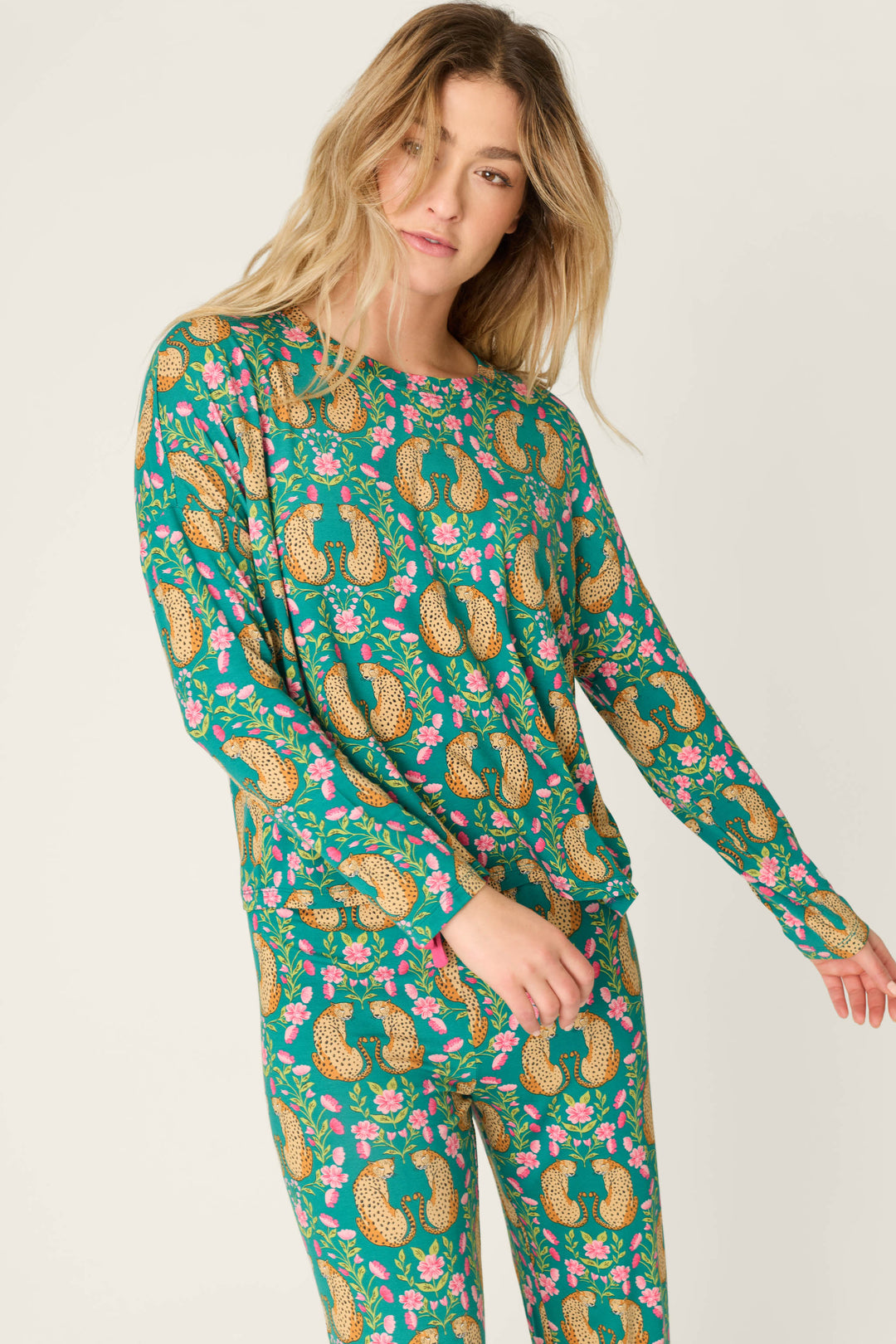 A woman wears the P.J. Salvage Cheetah Bonita Dreams Cheetah Print Long Sleeve Top, featuring a vibrant pattern of leopards and pink flowers, as she stands against a neutral background.