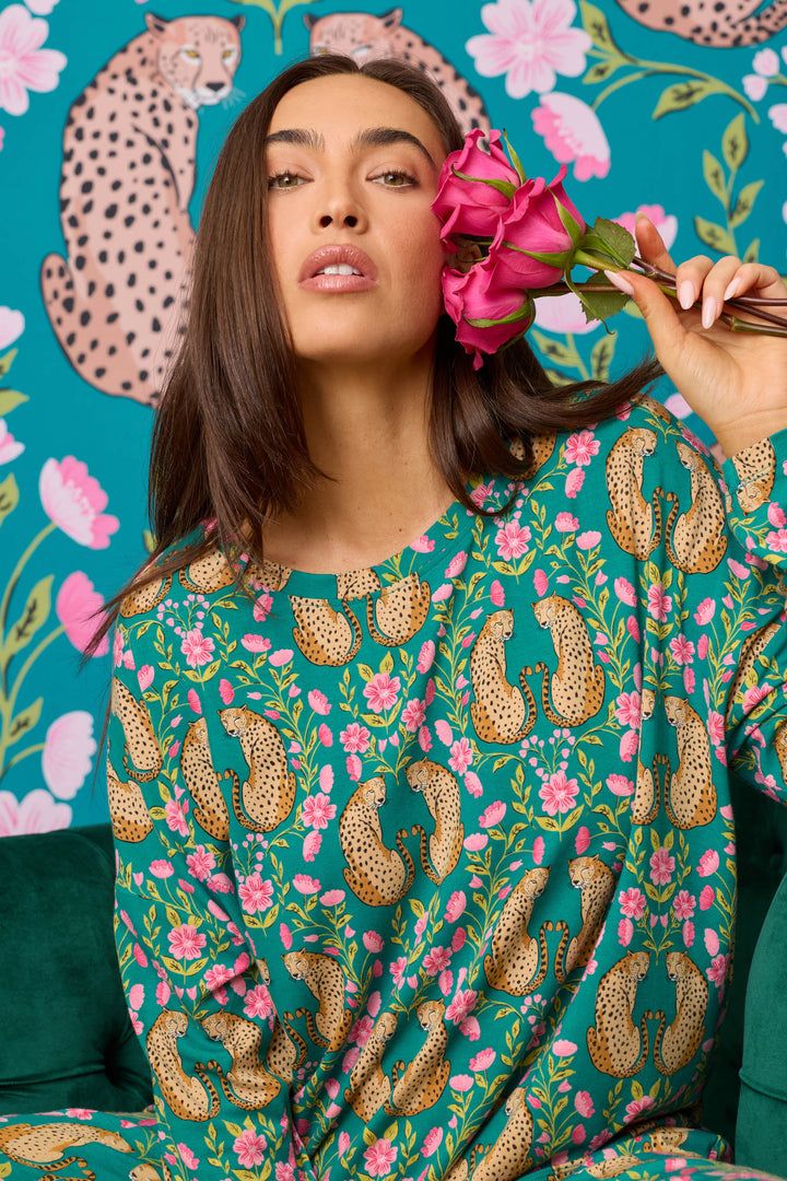 Wearing the P.J. Salvage Cheetah Bonita Dreams Cheetah Print Long Sleeve Top, a woman holds pink roses near her face while sitting in front of coordinating cheetah and floral wallpaper.