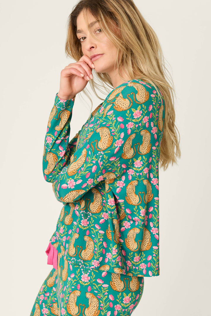 A woman with long blonde hair is wearing the P.J. Salvage Cheetah Bonita Dreams Cheetah Print Long Sleeve Top in a matching green set featuring cheetah and floral patterns, standing in profile with her hand touching her chin and looking at the camera.