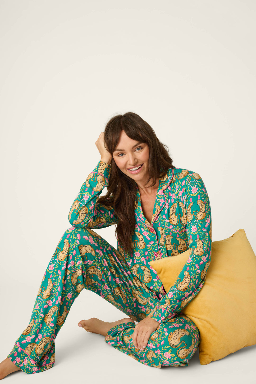 Wearing the P.J. Salvage Cheetah Bonita Dreams Cheetah Print Long Sleeve PJ Set, a woman sits on the floor, smiling and leaning on a yellow pillow against a plain background.