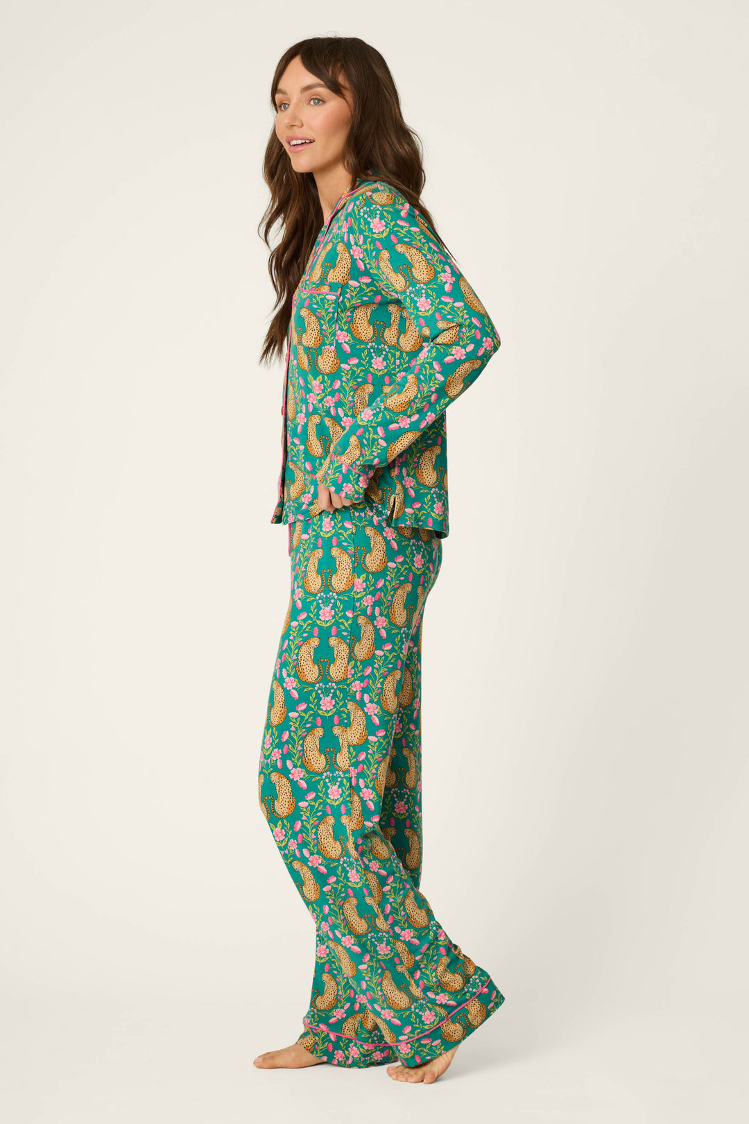 A woman stands sideways, barefoot, wearing the P.J. Salvage Cheetah Bonita Dreams Cheetah Print Long Sleeve PJ Set, which features a colorful cheetah pattern on green fabric. The pajama set includes a long-sleeve button-up top and loose pants. She has long brown hair and is smiling slightly.