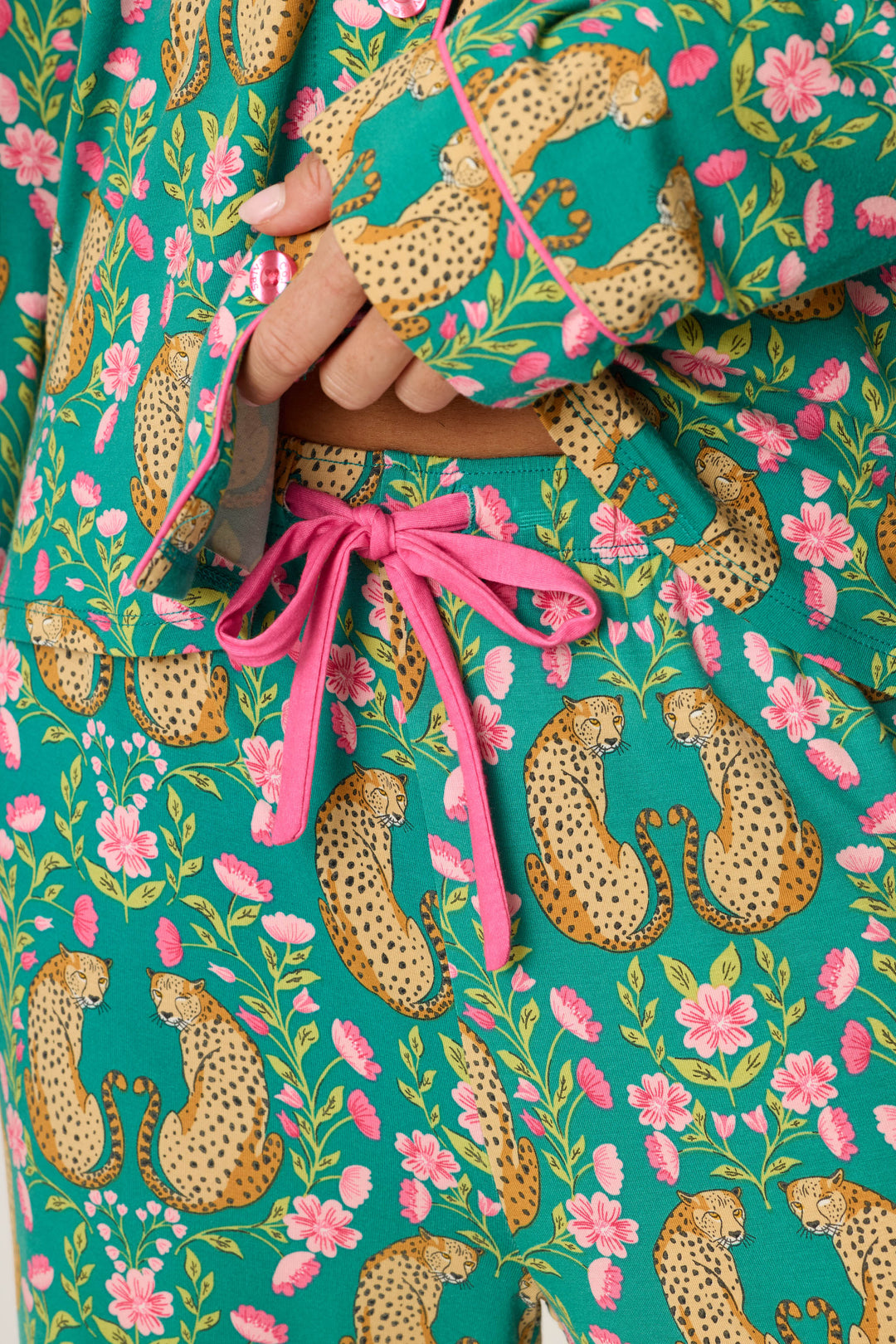 A close-up of a person wearing the P.J. Salvage Cheetah Bonita Dreams Cheetah Print Long Sleeve PJ Set shows teal pajamas with a pink drawstring, adorned with cheetahs and pink flowers. The person’s hand rests near the waistband.