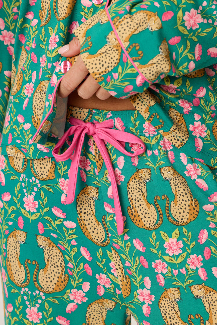 A close-up of a person wearing the P.J. Salvage Cheetah Bonita Dreams Cheetah Print Long Sleeve PJ Set shows teal pajamas with a pink drawstring, adorned with cheetahs and pink flowers. The person’s hand rests near the waistband.
