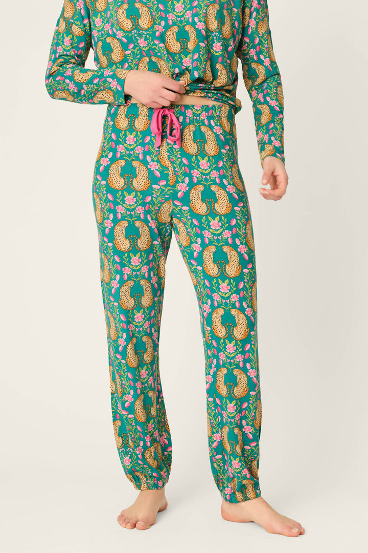 A person wearing P.J. Salvage Cheetah Bonita Dreams Cheetah Print Pajama Joggers, featuring a colorful cheetah pattern, stands barefoot against a plain background. Only the lower body and part of the upper body are visible.