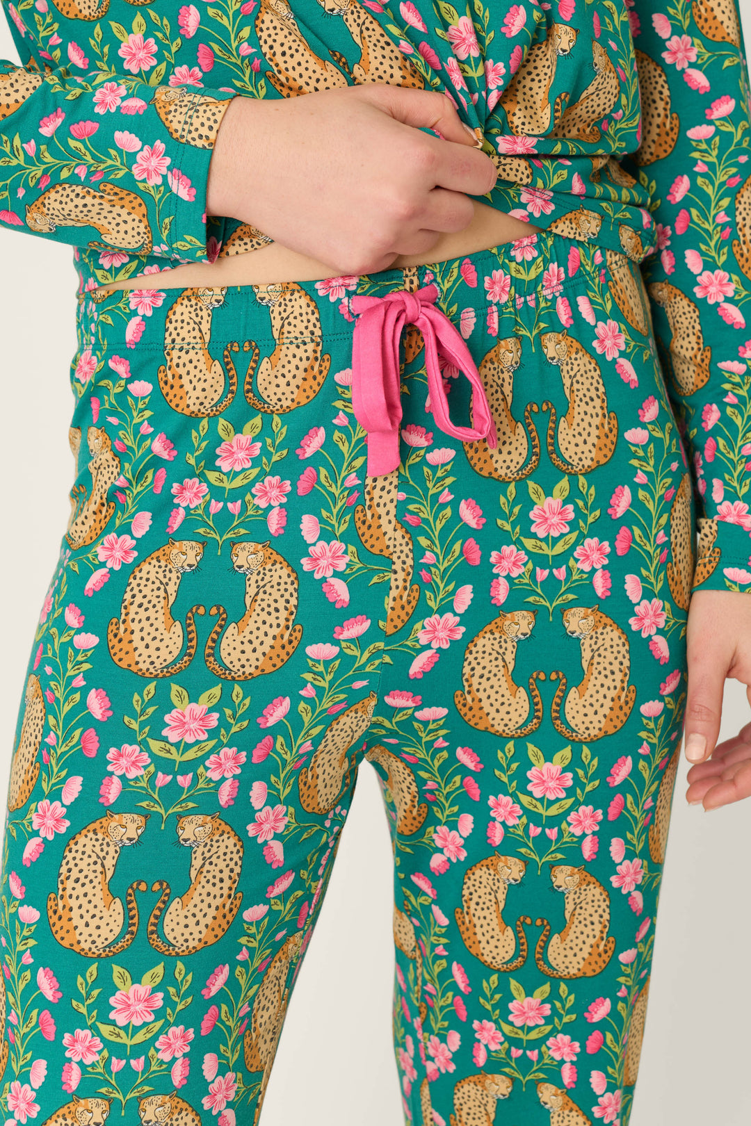 Model is wearing the P.J. Salvage Cheetah Bonita Dreams Cheetah Print Pajama Joggers, featuring a vibrant mix of cheetah, floral, and leaf patterns on a teal background with pink drawstring ties at the waist. Only the torso and upper legs are shown.