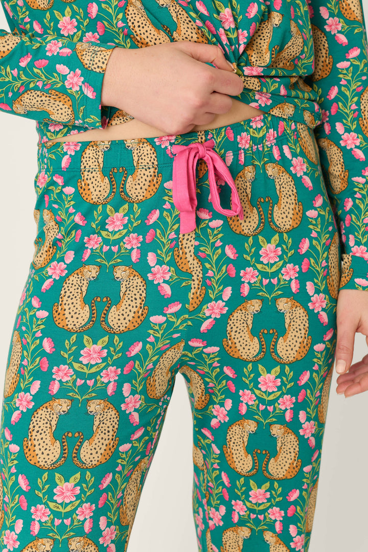 Model is wearing the P.J. Salvage Cheetah Bonita Dreams Cheetah Print Pajama Joggers, featuring a vibrant mix of cheetah, floral, and leaf patterns on a teal background with pink drawstring ties at the waist. Only the torso and upper legs are shown.