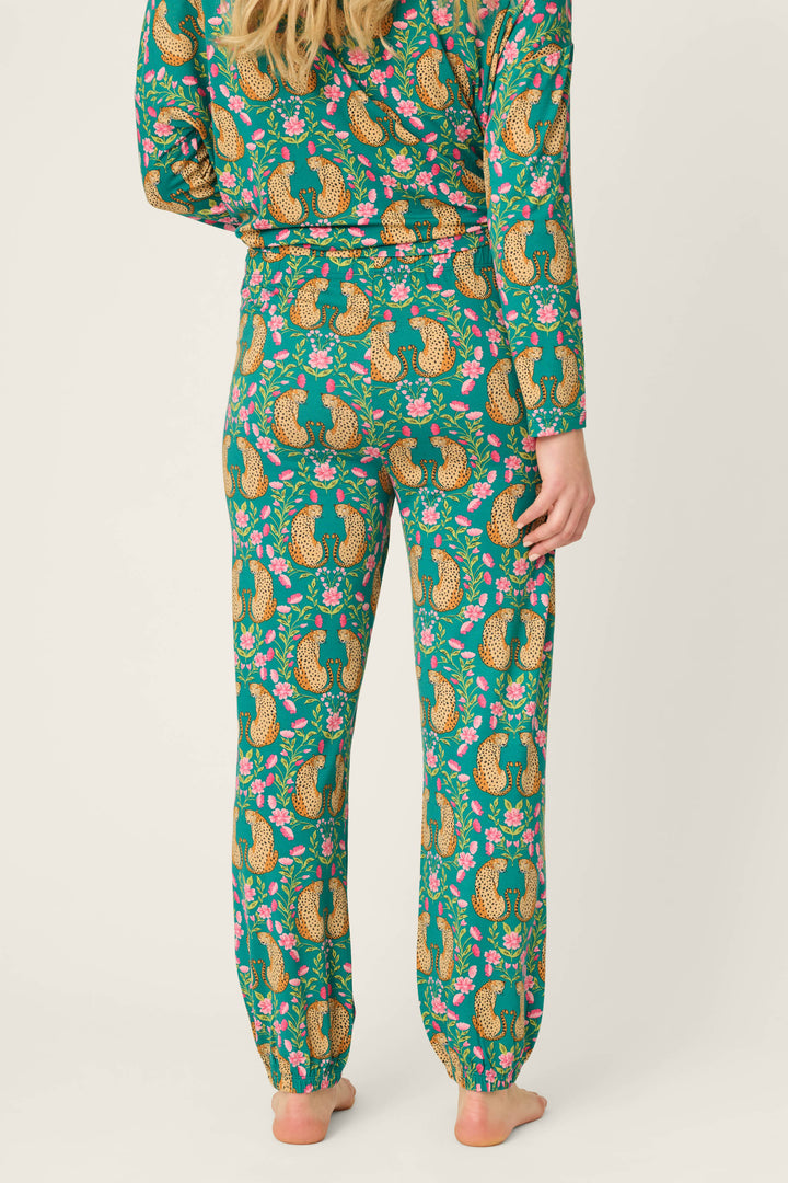 A person stands barefoot, shown from the back, wearing P.J. Salvage's Cheetah Bonita Dreams Cheetah Print Pajama Joggers featuring a colorful green pattern with cheetahs and pink flowers; the matching set includes a long-sleeve top and full-length pants.