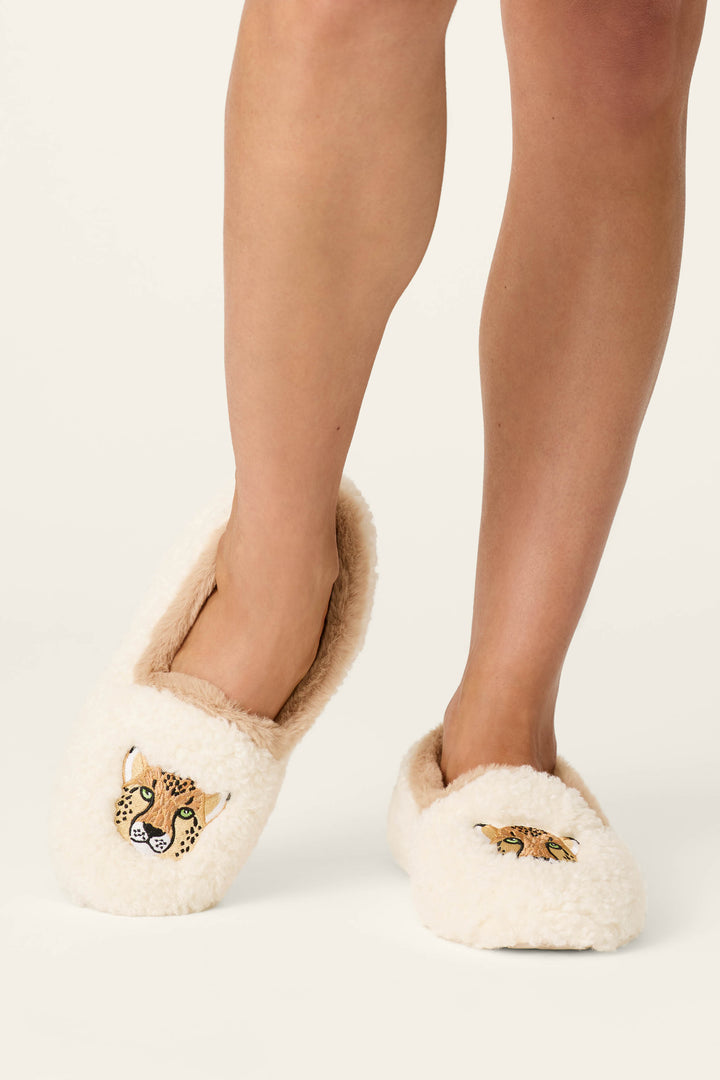 A person wearing P.J. Salvage Cheetah Bonita Dreams Embroidered Cheetah Slippers, featuring a soft, cozy cream-colored design with cartoon cheetah faces on each slipper. The person's lower legs are visible against a plain background.