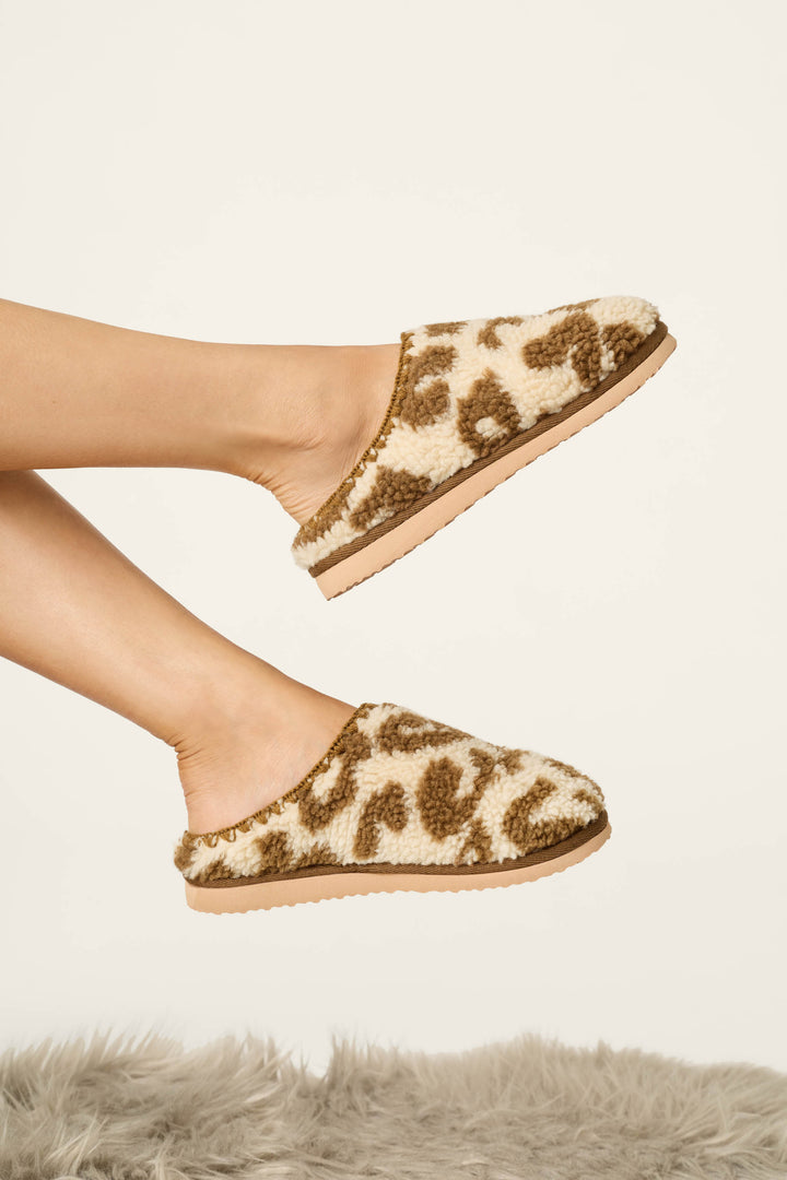 A person’s feet wearing P.J. Salvage Cheetah Bonita Dreams Cheetah Print Fuzzy Slippers, hovering above a soft, gray shaggy rug on a neutral background.