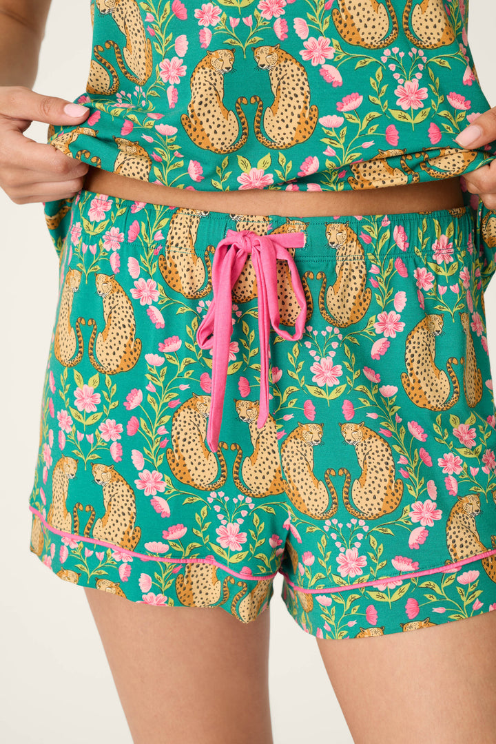Wearing the P.J. Salvage Cheetah Bonita Dreams Cheetah Print Pajama Shorts, this person pairs the green cheetah and pink flower print shorts with a matching top, lifting the shirt slightly to reveal the pink drawstring and waistband details.