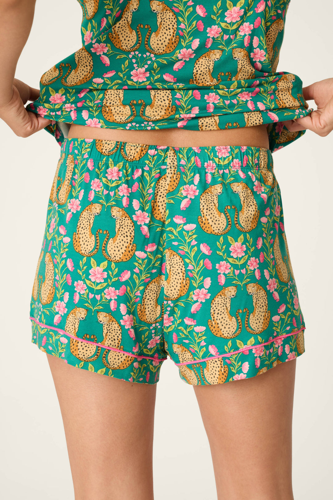 A person wearing matching green P.J. Salvage Cheetah Bonita Dreams cheetah print pajama shorts and top, featuring a leopard and pink flower pattern, lifts the shirt slightly to reveal the waistband of the shorts. The background is plain and light-colored.
