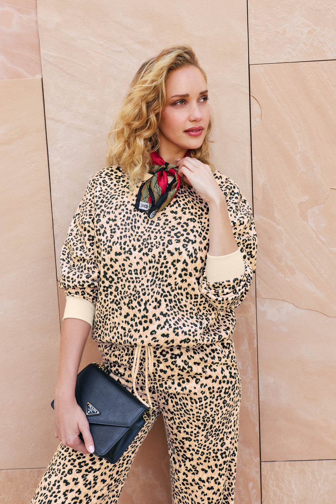 A woman in the C'est La Vie Leopard Print Frankie Sweater by P.J. Salvage poses against a beige stone wall, holding a black Prada clutch and wearing a red patterned scarf around her neck. She has wavy blonde hair and looks off to the side, smiling slightly.