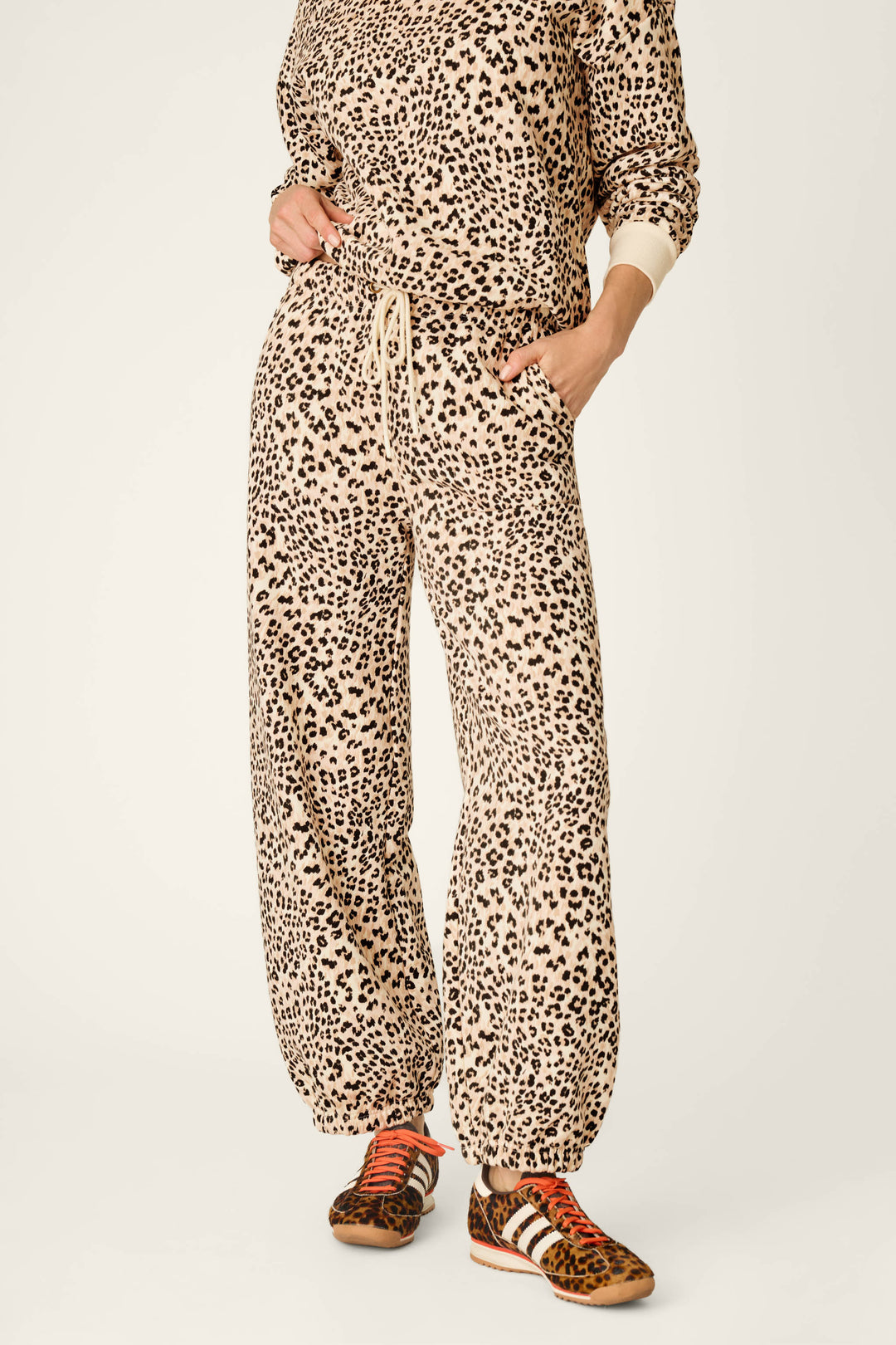 A person wearing the P.J. Salvage C'est La Vie Leopard Print Good Hang Jogger Pants, styled with a matching beige and black leopard-print sweatshirt and brown leopard-print sneakers with red laces, hands in pockets and face not visible.