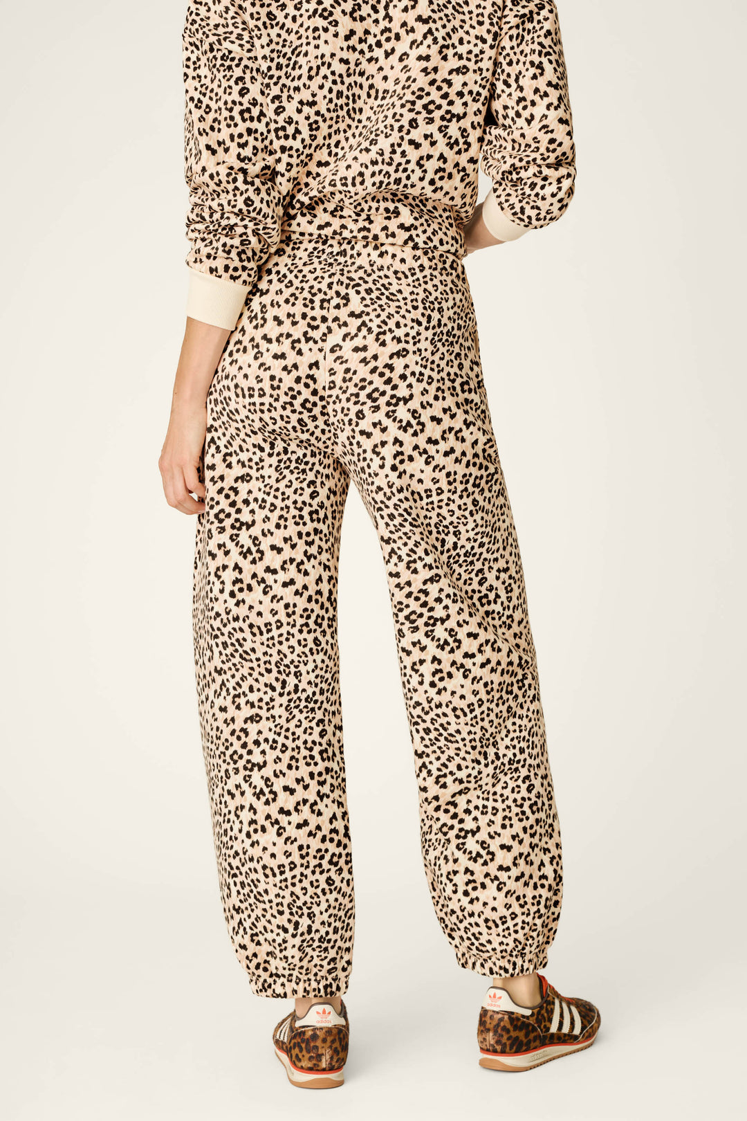 A person stands facing away, wearing the P.J. Salvage C'est La Vie Leopard Print Good Hang Jogger Pants in a beige and black leopard print set, paired with brown sandals. The background is plain and light-colored.