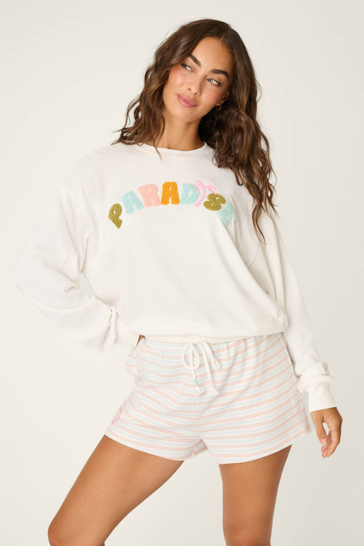A woman models the P.J. Salvage Cheeky Paradise Sweatshirt, featuring colorful "PARADISE" lettering, paired with light striped shorts and posing with one hand on her hip, gazing to the side.
