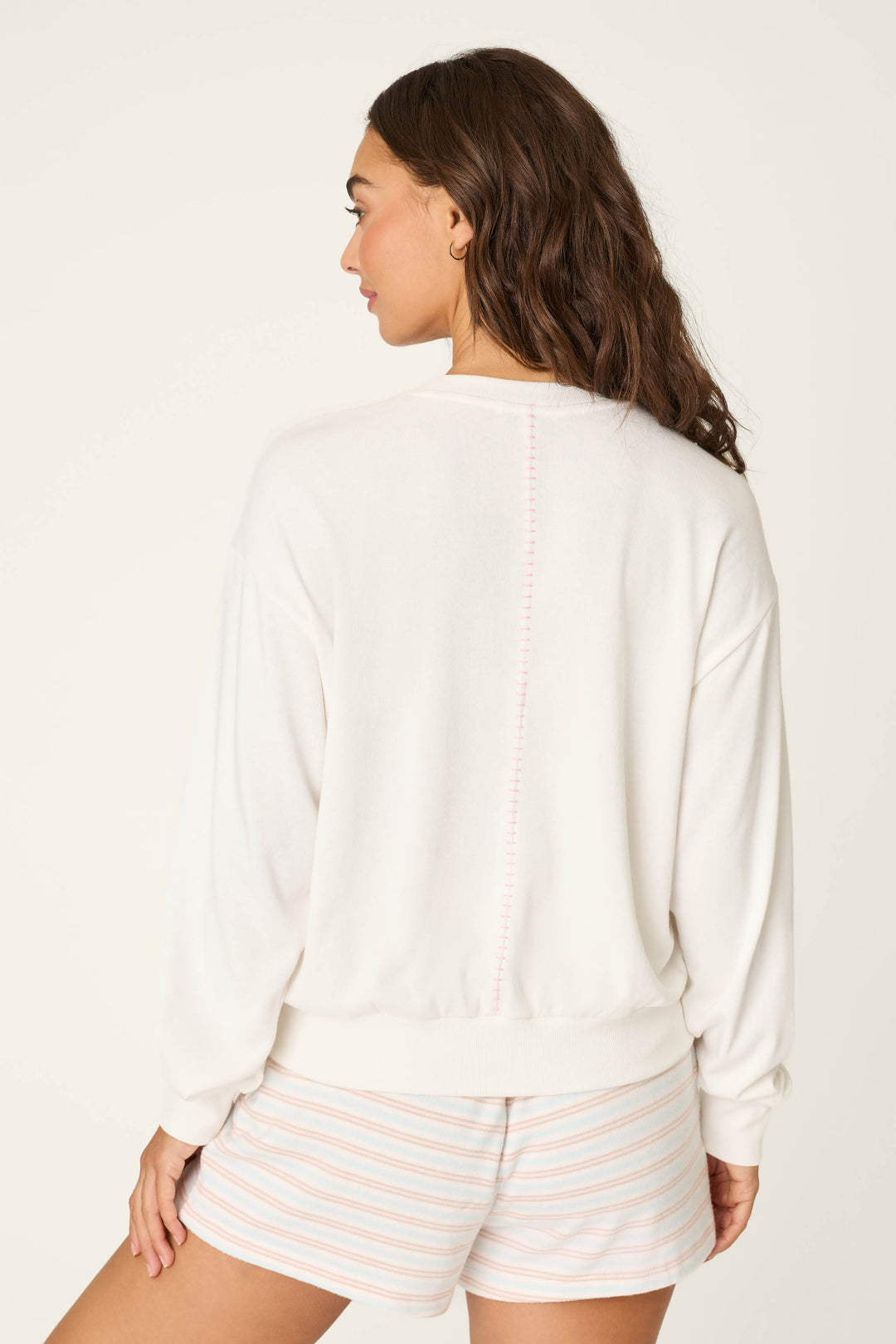 A woman with wavy brown hair faces away, wearing the P.J. Salvage Cheeky Paradise Sweatshirt—a white long-sleeve top with subtle pink back stitching—and light shorts with pink stripes, set against a plain light background.