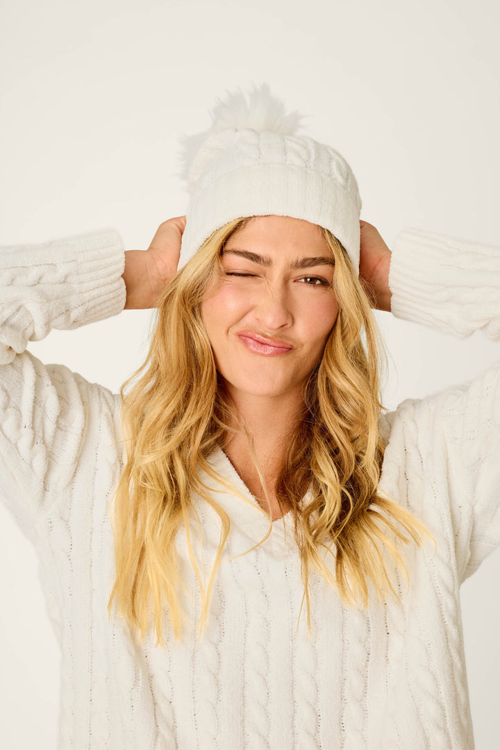 A woman with long blonde hair, playfully winking and smiling, presses her hands to her P.J. Salvage Cozy Chenille Cable Knit Ivory Beanie with Pompom, paired with a white sweater against a plain background.
