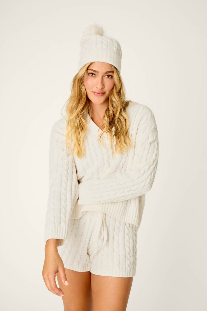 A woman with long blonde hair stands against a plain light background, wearing a white knit sweater and matching knit shorts, styled with the P.J. Salvage Cozy Chenille Cable Knit Ivory Beanie with Pompom.