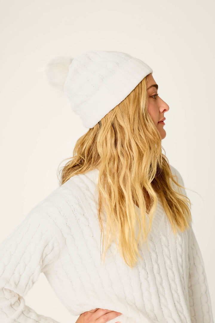 A woman wearing a Cozy Chenille Cable Knit Ivory Beanie with Pompom by P.J. Salvage stands sideways with her hand on her hip, looking off to the right against a plain light background.