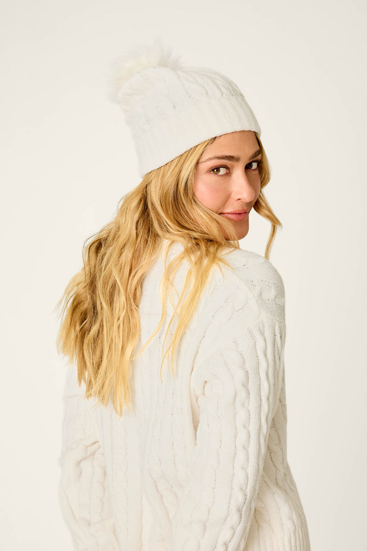 A person with long blonde hair wears the P.J. Salvage Cozy Chenille Cable Knit Ivory Beanie with Pompom and a matching white knit sweater, looking over their shoulder and smiling softly against a light background.