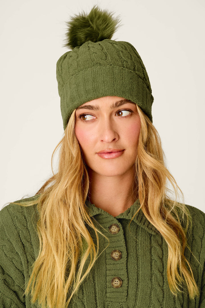 A woman with long blonde hair wears a green knit sweater and the P.J. Salvage Cozy Chenille Cable Knit Olivine Beanie with Pompom, looking slightly to the side.