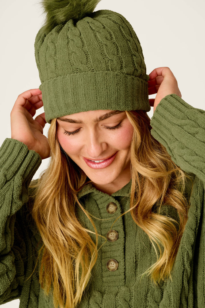 A woman smiles with her eyes closed as she adjusts the Cozy Chenille Cable Knit Olivine Beanie with Pompom by P.J. Salvage, paired with a green knit sweater featuring three buttons.