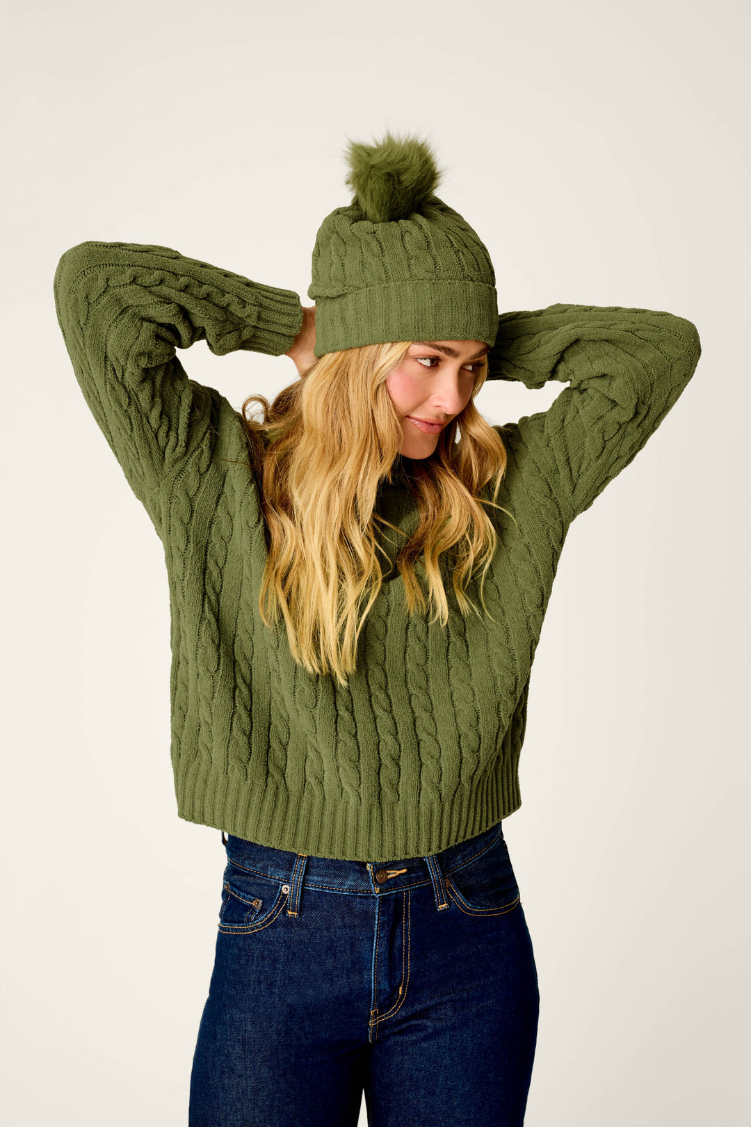 A woman with long blonde hair smiles softly as she stands against a plain background, adjusting her P.J. Salvage Cozy Chenille Cable Knit Olivine Beanie with Pompom.