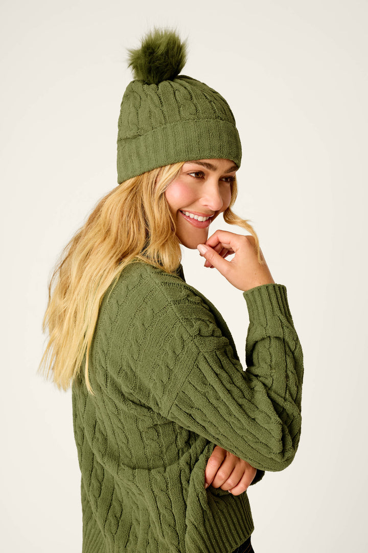 A woman smiles while looking to the side against a plain background, wearing the P.J. Salvage Cozy Chenille Cable Knit Olivine Beanie with Pompom and a matching green sweater.